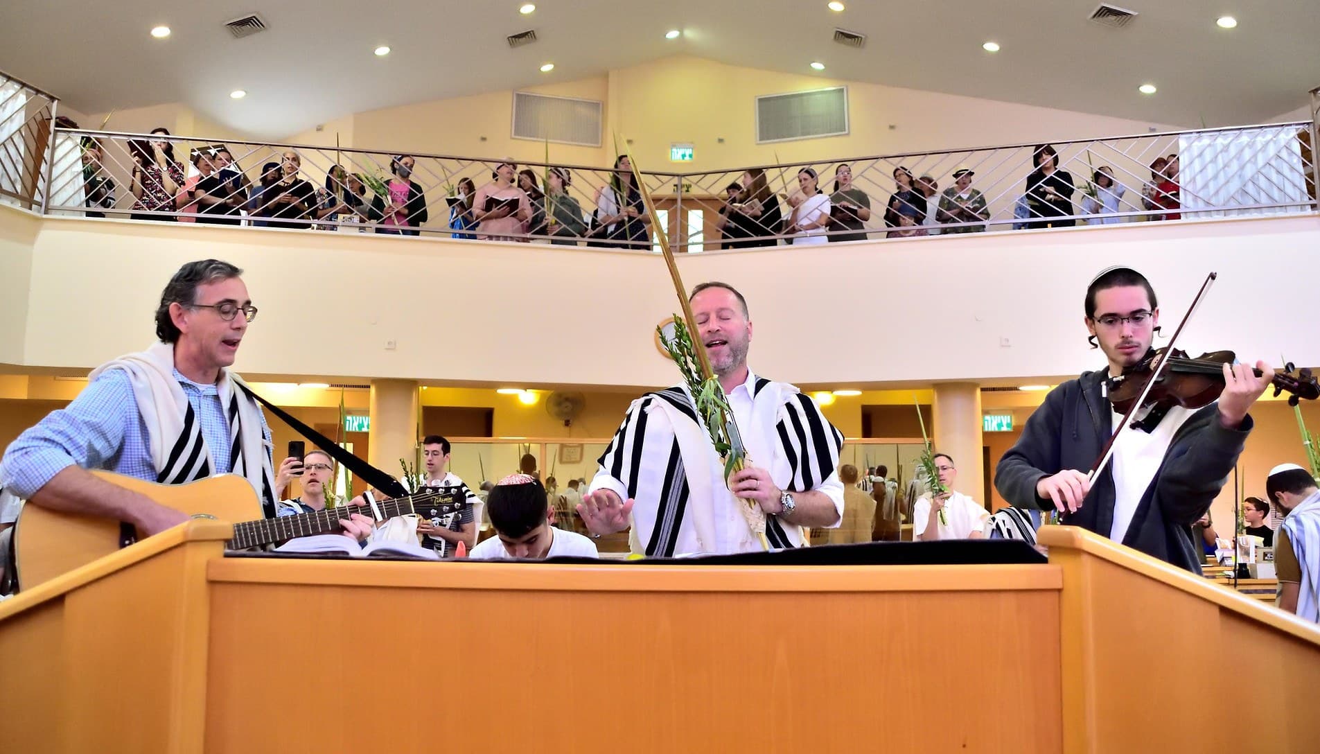 Hallel on Sukkot at Ohel Yona Menachem in Beit Shemesh: men in tallitot with lulavim at the bimah, one playing guitar and one the violin, the women's gallery full above