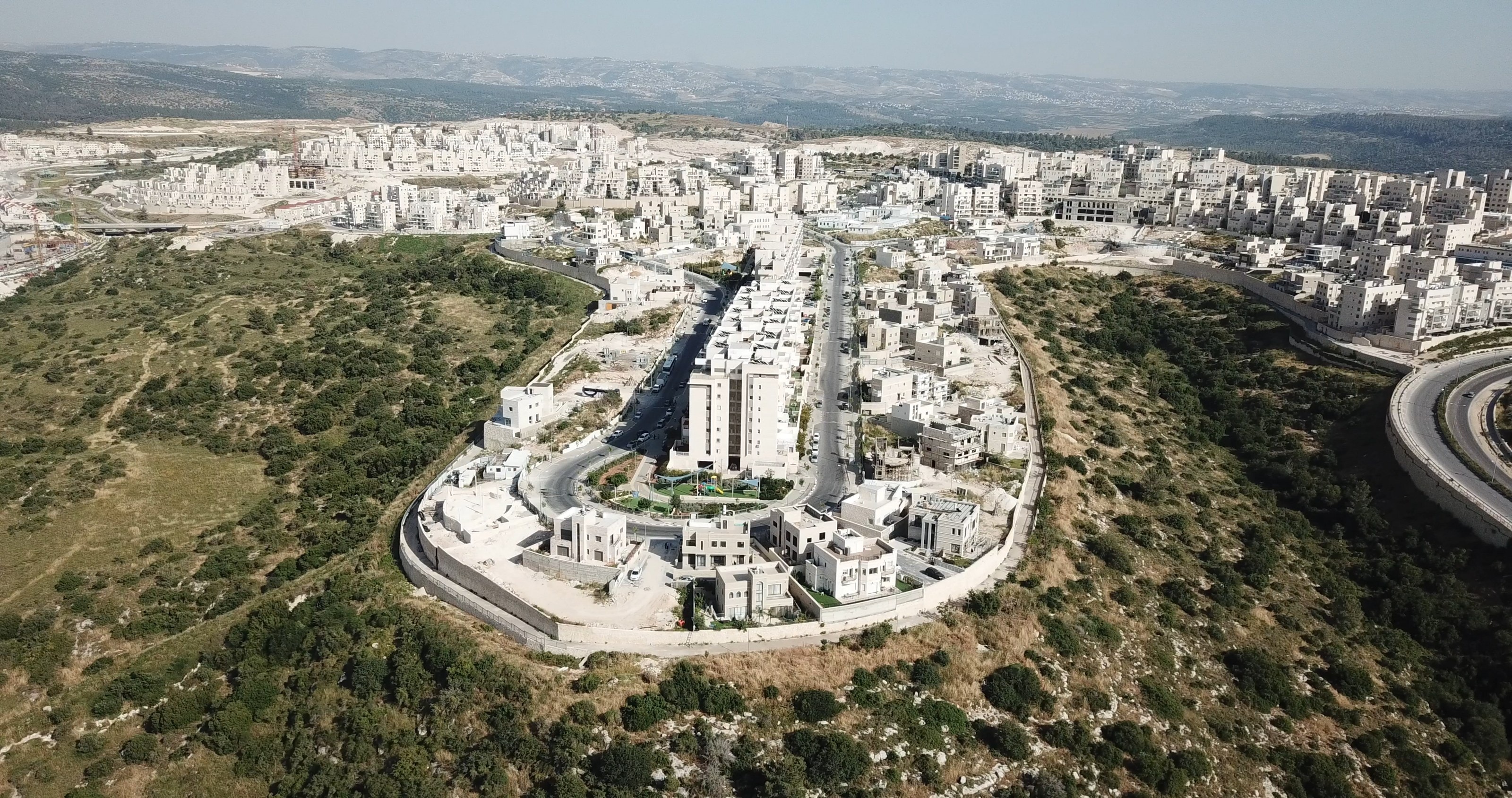 Aerial view of Ramat Beit Shemesh Gimmel: white stone apartment blocks along curving streets on a ridge, wooded hills around