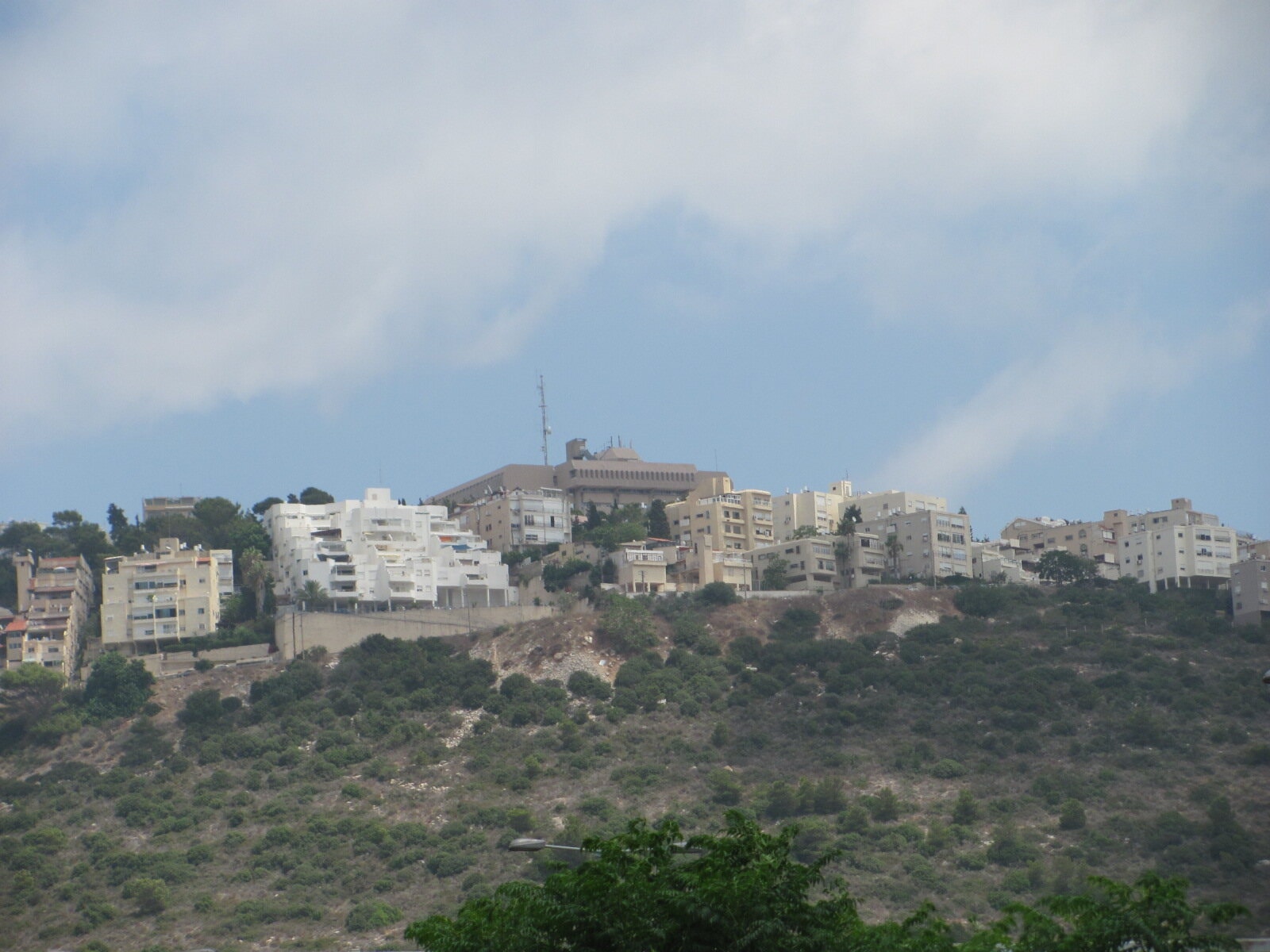 Apartment blocks in pale stone along the top of a green, scrubby ridge under a cloudy sky