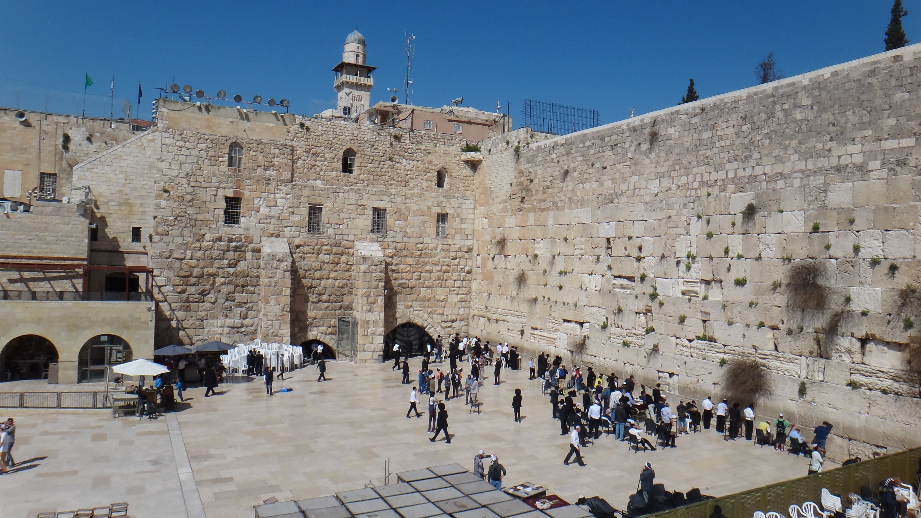 The Western Wall rising over its plaza under a blue sky, people praying at its foot