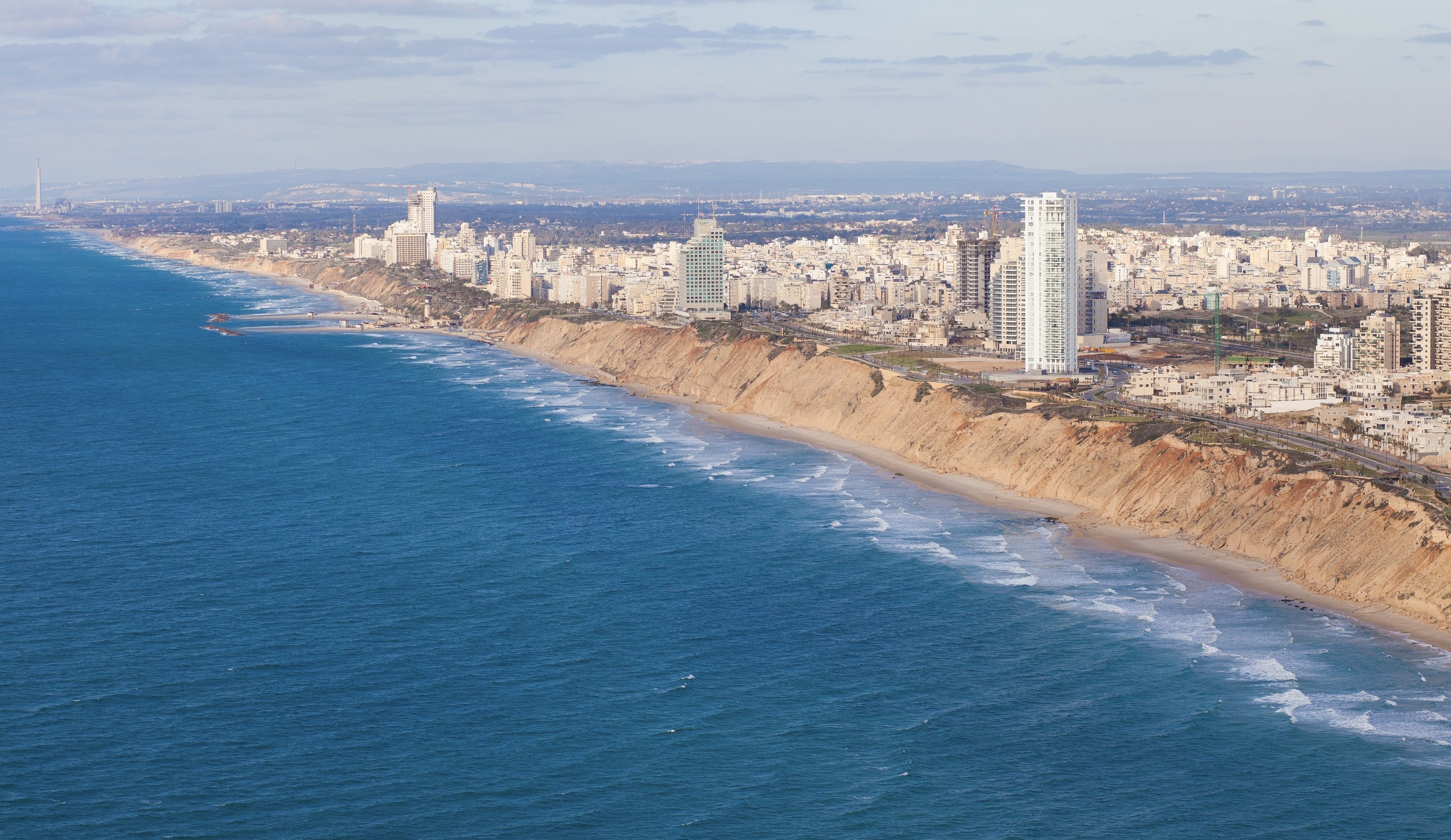 Netanya from the air in low sun: white apartment towers and hotels along the top of sandstone cliffs, with the beach and the open sea below