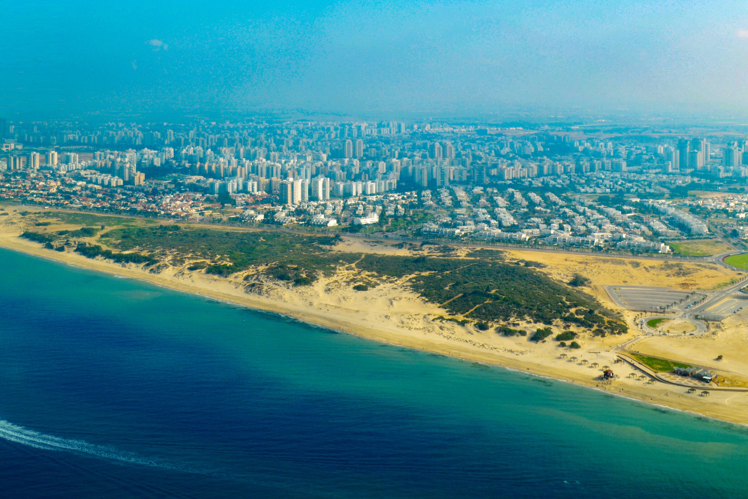 Ashdod from the air over the sea: a wide sandy beach and green dunes in front, rows of apartment towers behind them