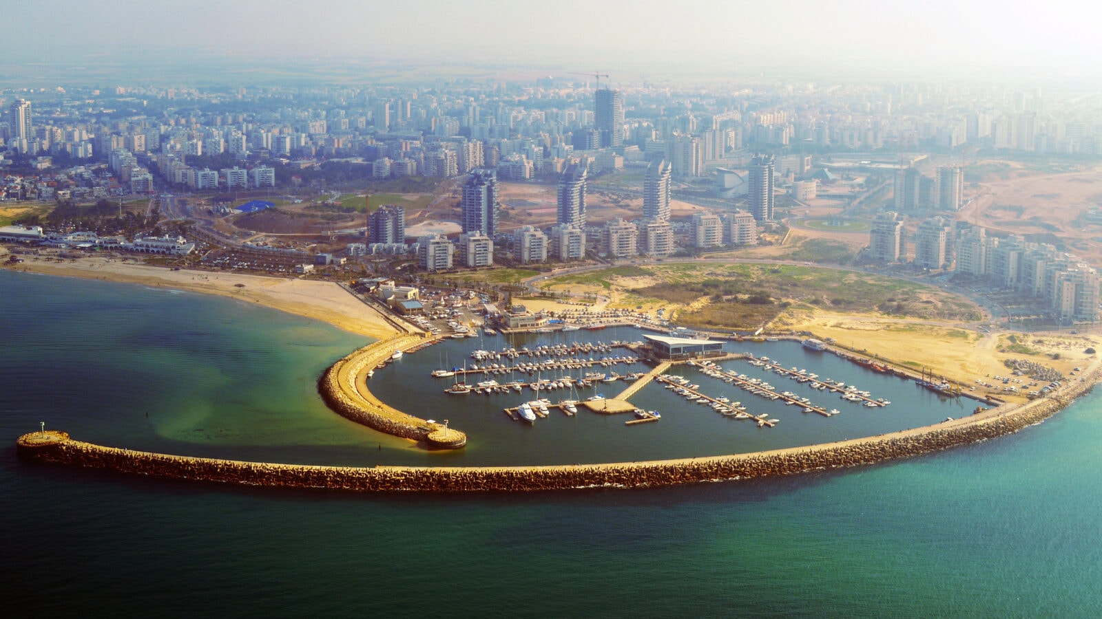 Ashdod Marina from the air: a curved breakwater around moored boats, with apartment towers and open sand behind