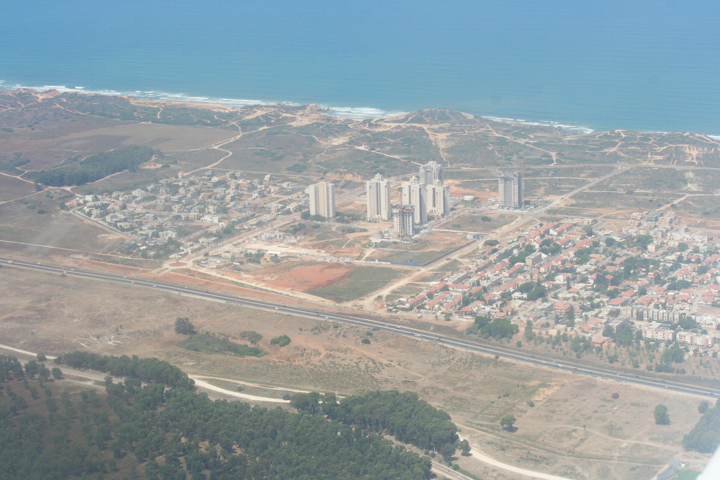 Givat Olga from the air: low houses with red roofs and a cluster of apartment towers between a highway and sand dunes, the sea behind