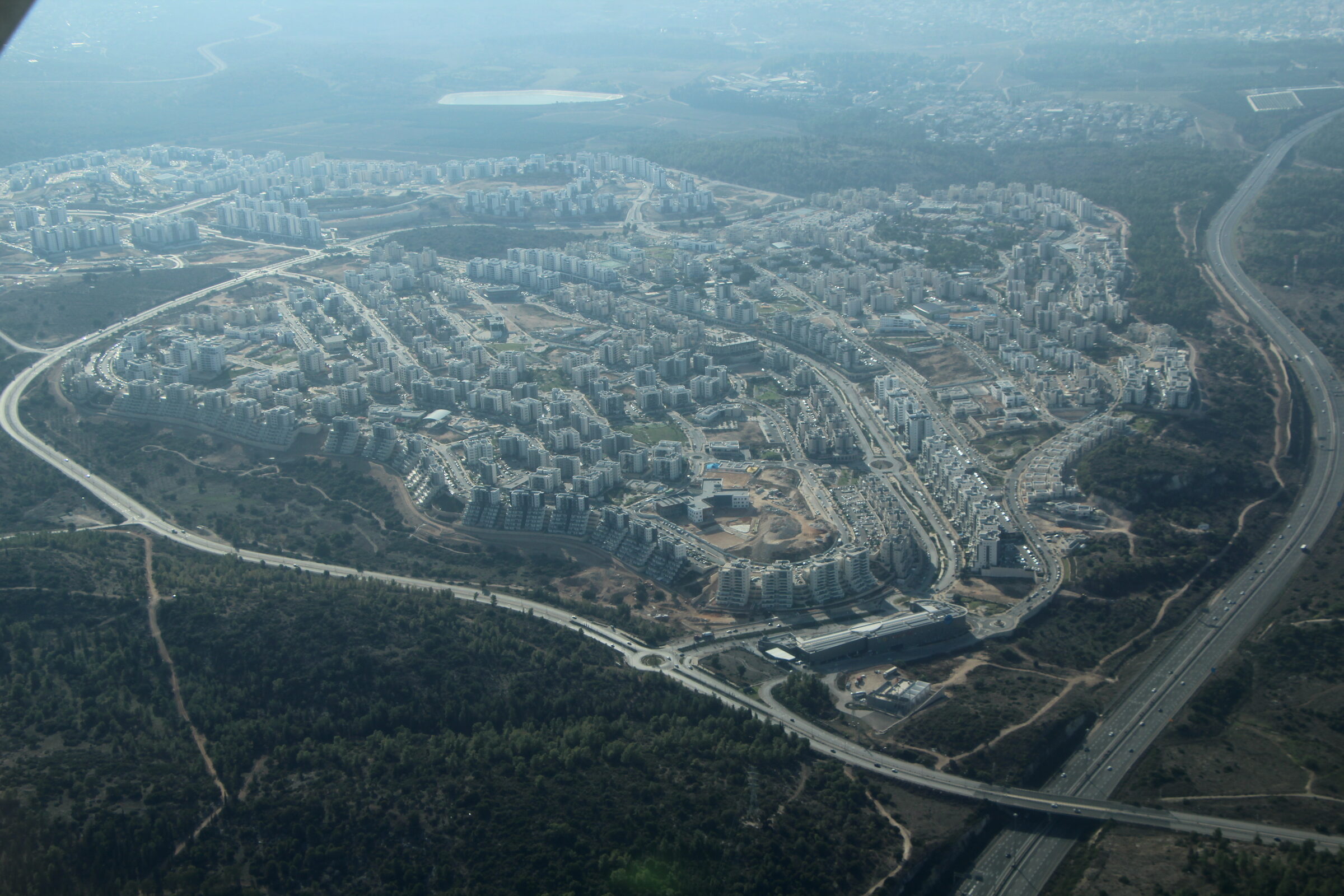 Harish from the air: rows of white apartment blocks on a rounded hill, ringed by a road and dark green forest, with Highway 6 running along the right edge