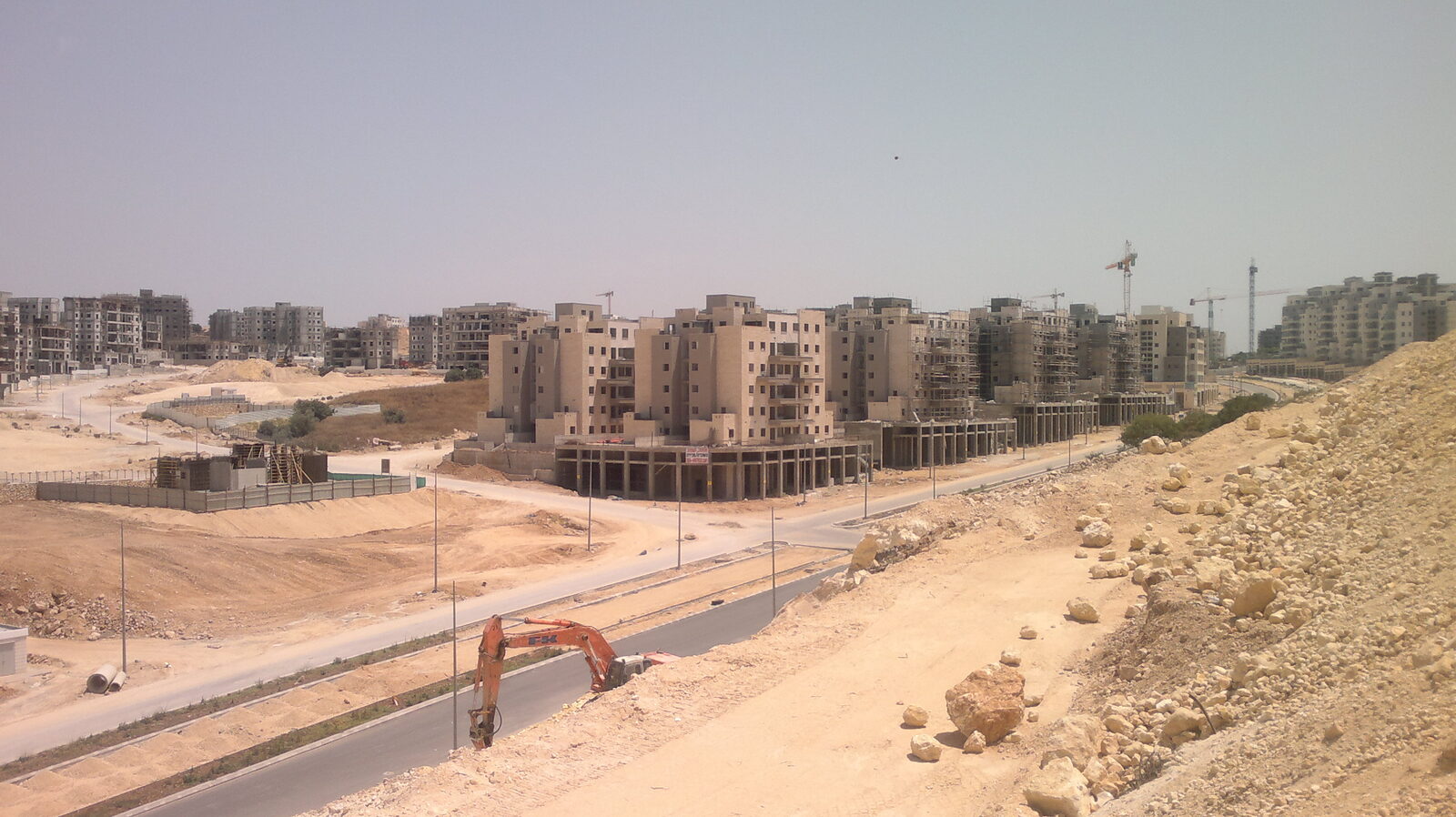 Half-built apartment buildings in pale stone on cleared sandy ground, with cranes behind and an excavator beside a new road