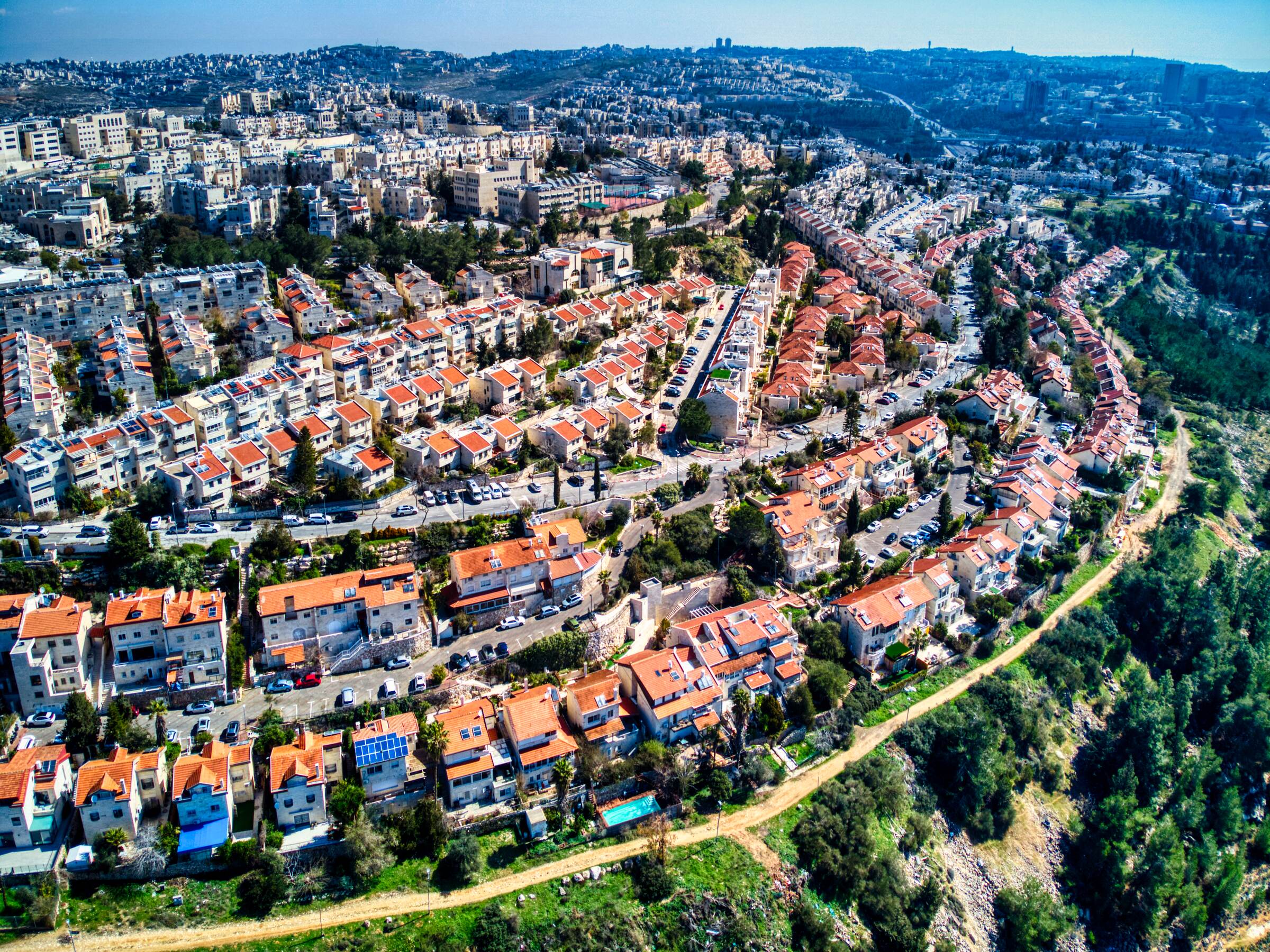 Rows of red-roofed terraced houses and apartment blocks on a hillside in Ramot, with a dirt path and pine trees along the slope