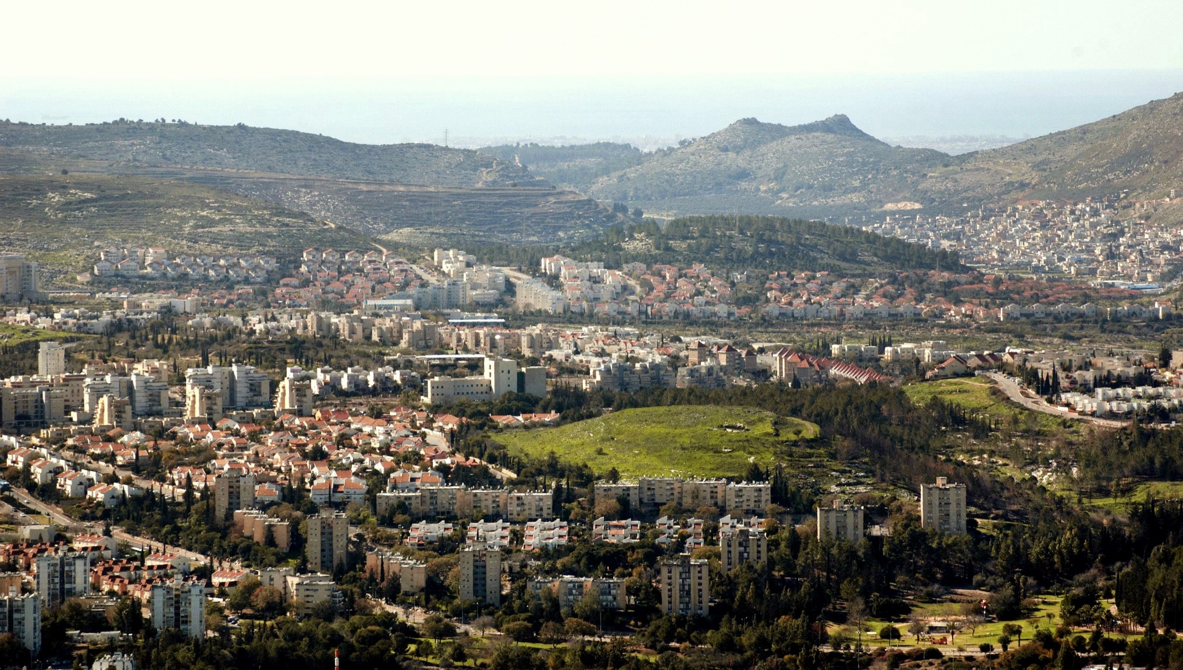 Karmiel seen from the hills to the east: apartment blocks and red-roofed houses spread across a green valley, with a grassy hill in the middle and ridges behind