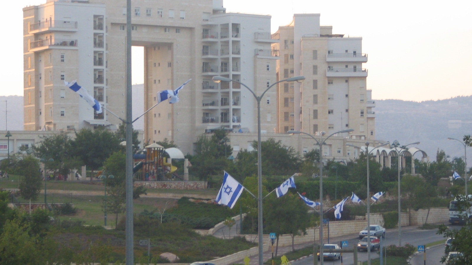 Tall pale stone apartment buildings in Ramat Rabin above a park with a playground, and a road lined with Israeli flags on lamp posts