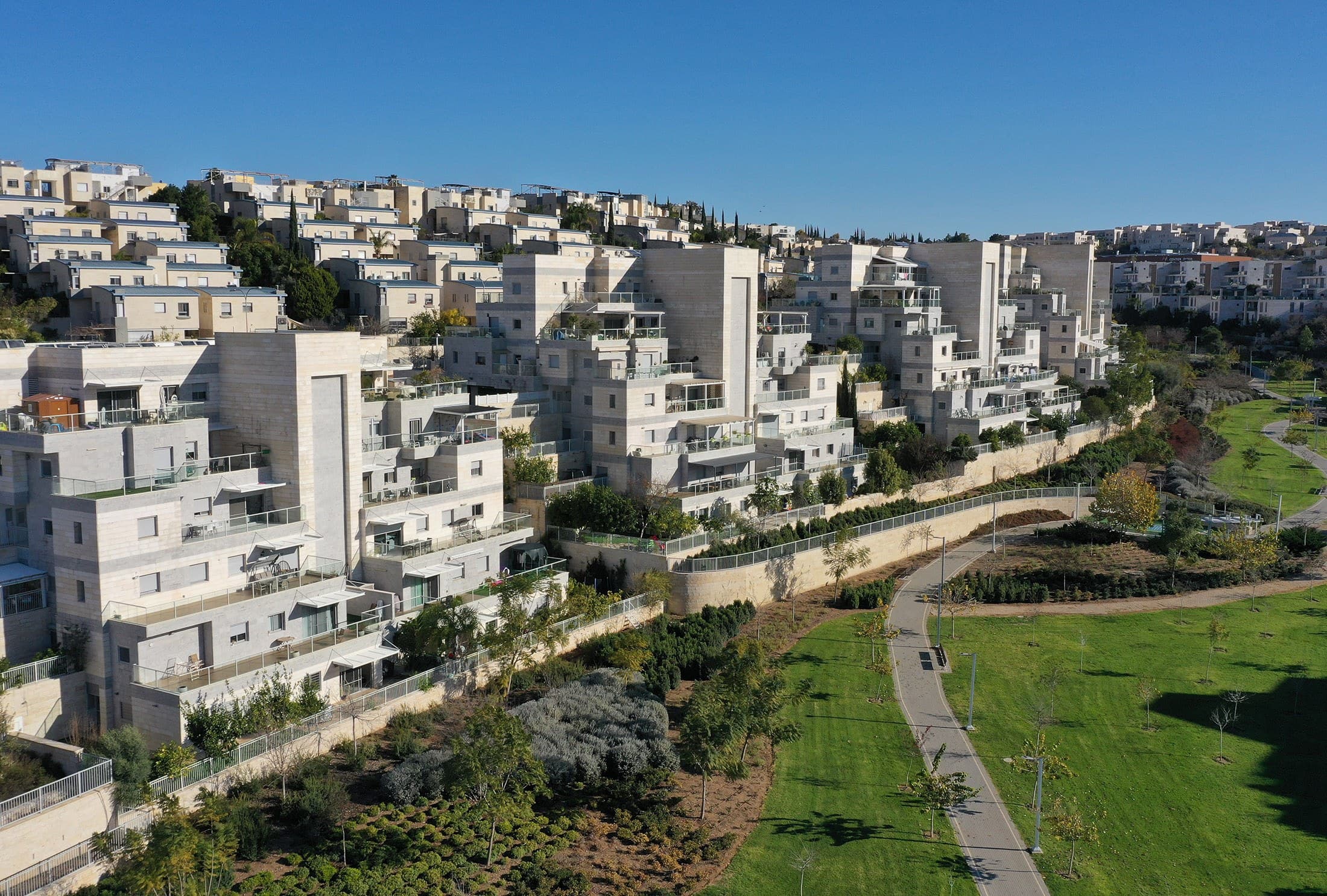A footpath through a lawn park below terraced stone apartment buildings in Buchman, Modi'in