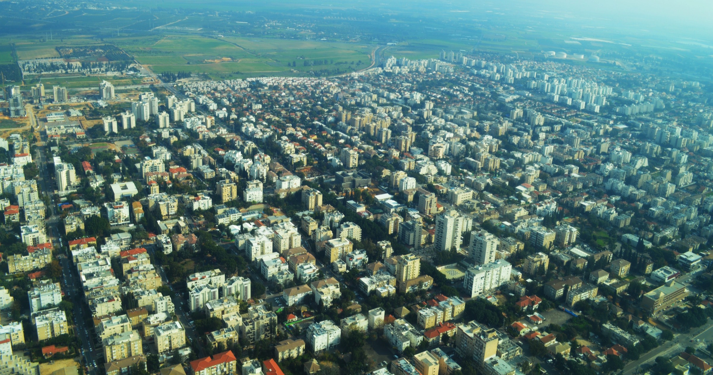 Rehovot from the air: streets of white apartment blocks and red-roofed houses among trees, with taller towers and open fields towards the horizon
