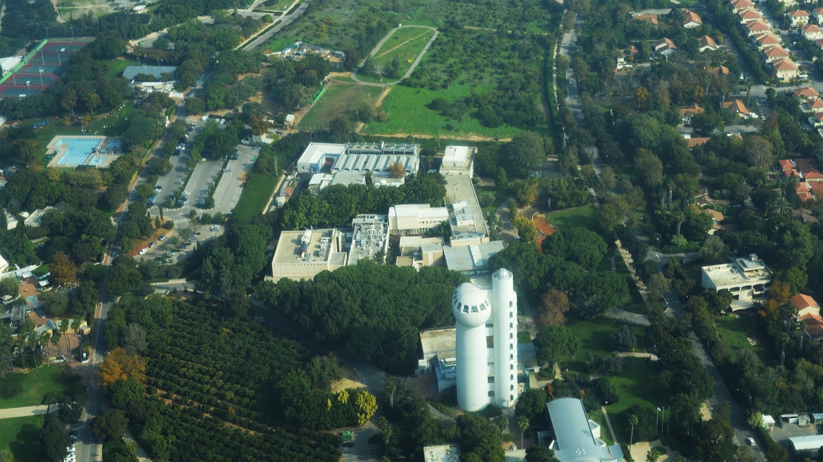 The Weizmann Institute of Science campus from the air: white laboratory buildings and a tall white tower among trees and orchards, with red-roofed houses along its eastern edge