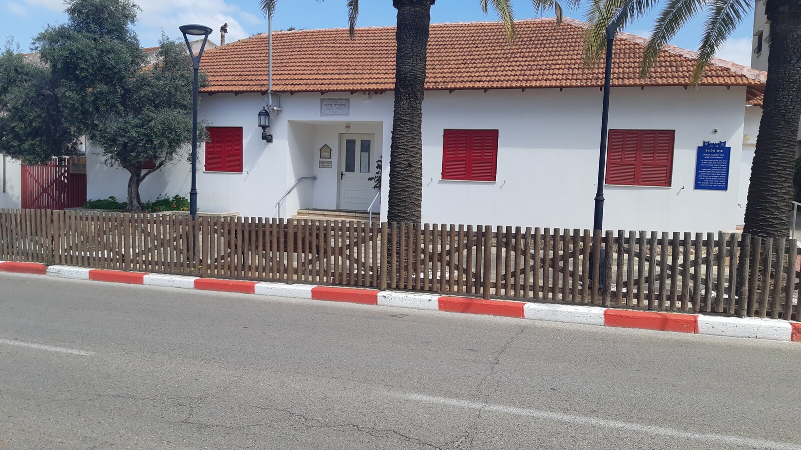 Beit HaLord: a single-storey white house with red shutters and a tiled roof behind a low wooden fence, with a blue heritage sign on the wall
