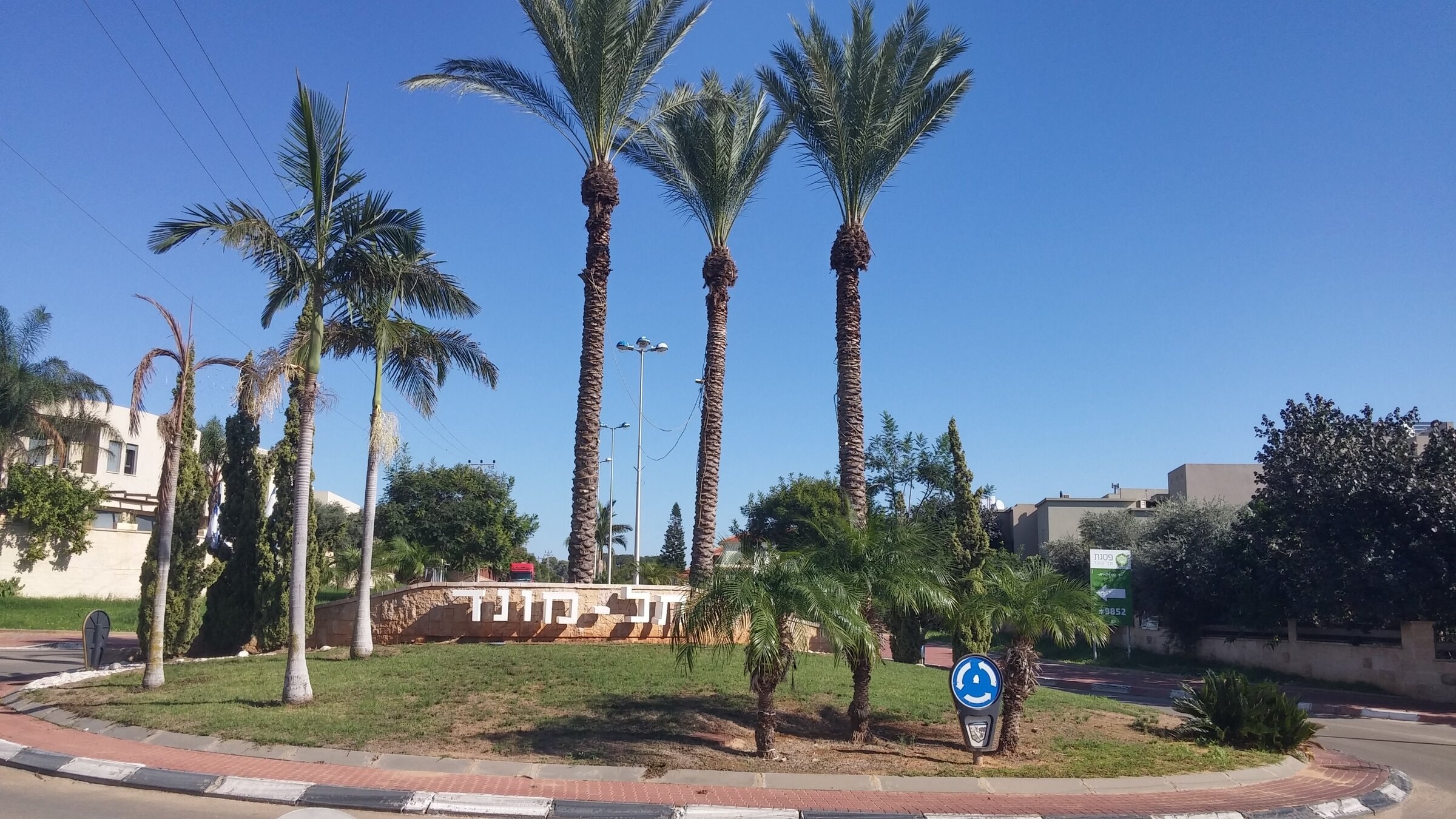 The entrance to Tel Mond: a grassy roundabout with tall date palms and a low stone wall carrying the town's name in Hebrew, with houses and trees behind