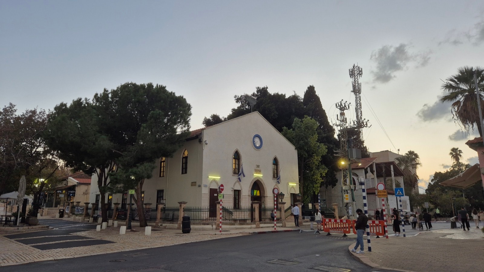 The Ohel Ya'akov Synagogue: a white stone building with arched windows and a round window under its gable, behind a low fence on a street corner at dusk