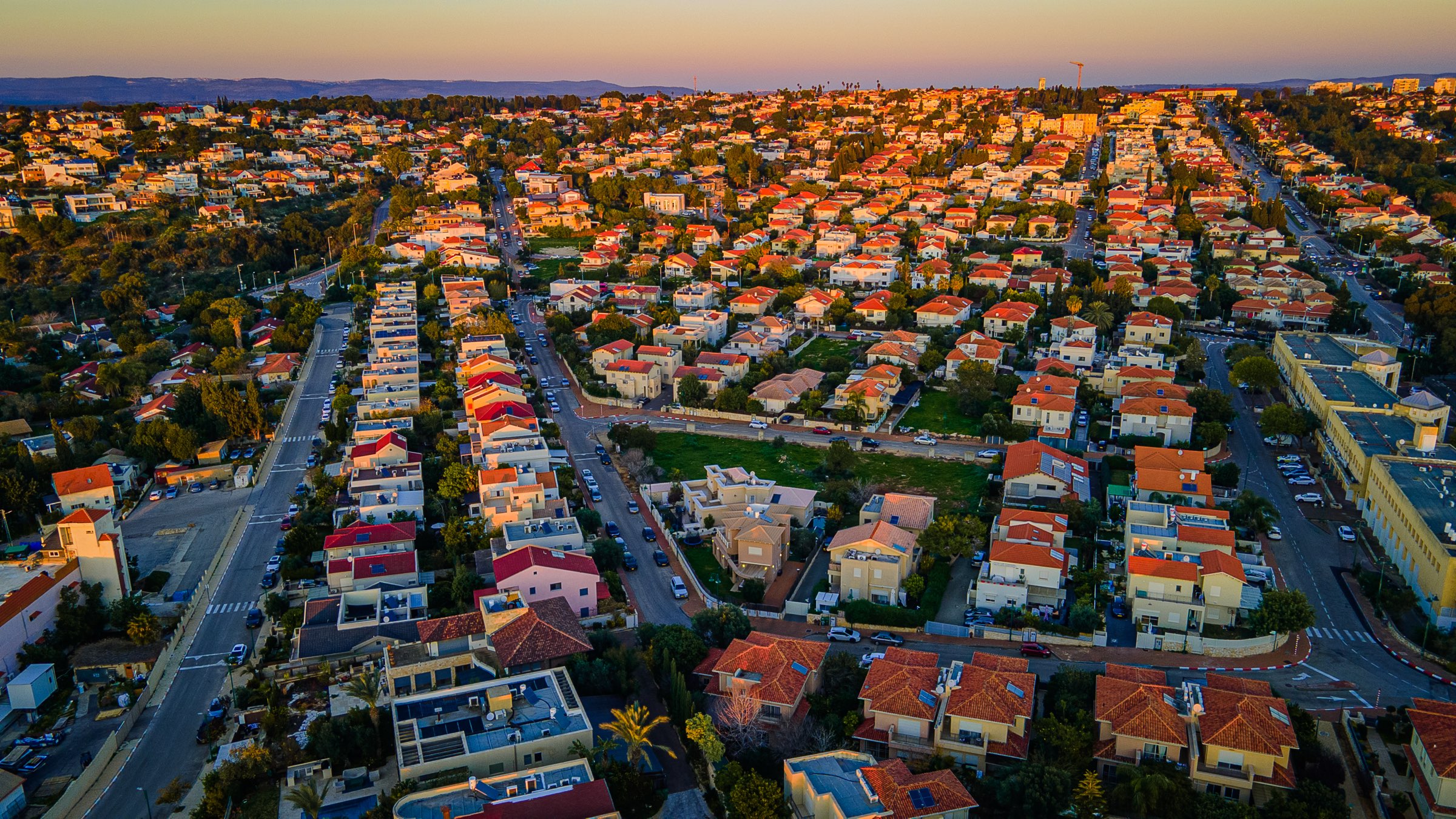 Zichron Yaakov from the air at sunset: streets of red-roofed houses and low apartment blocks among trees, with the Carmel hills on the horizon